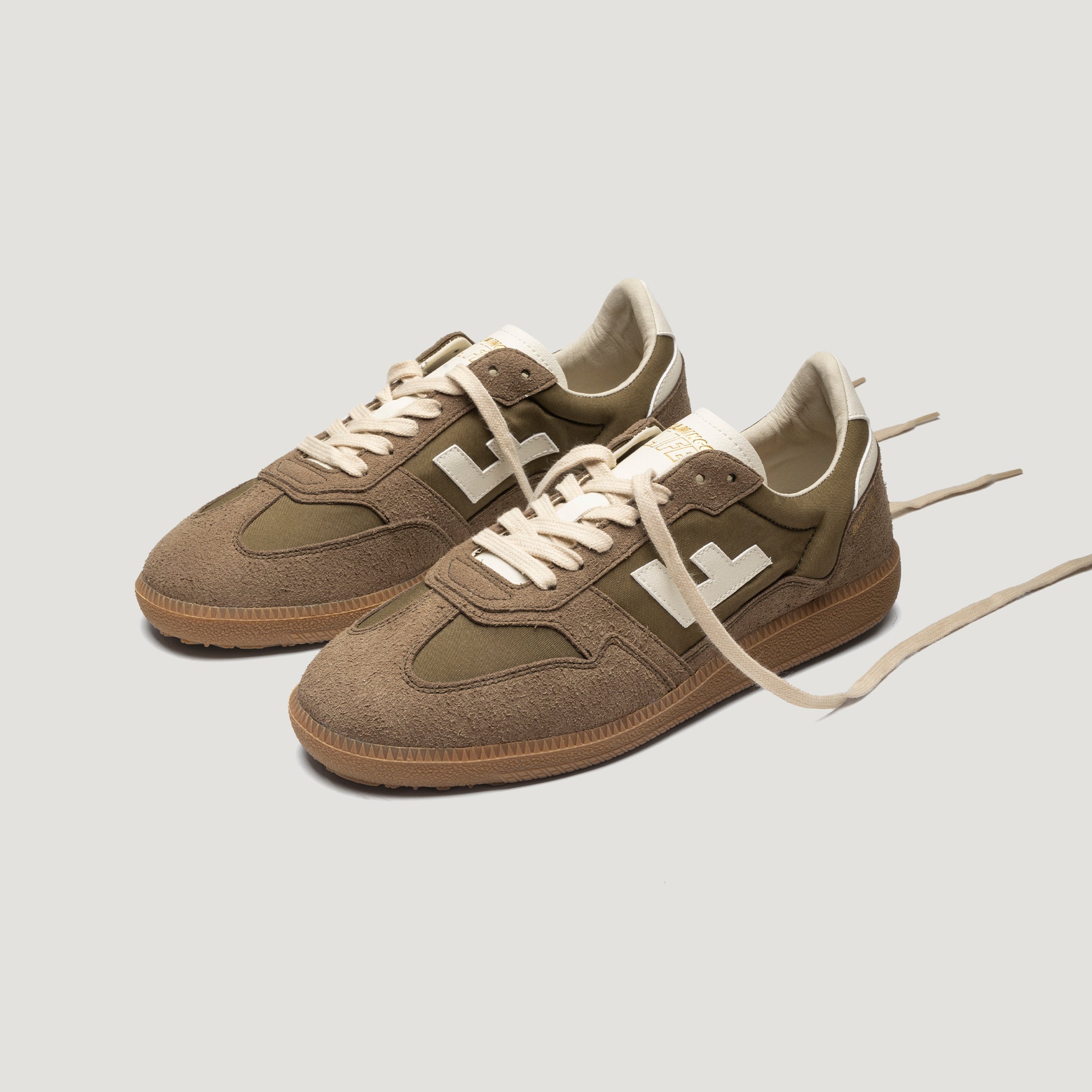 Ein Paar braune Wildleder-Turnschuhe mit weißen Schnürsenkeln und einem weißen "H"-Logo an der Seite auf einem hellgrauen Hintergrund. Die Schuhe sind schräg gestellt, die Schnürsenkel sind offen.