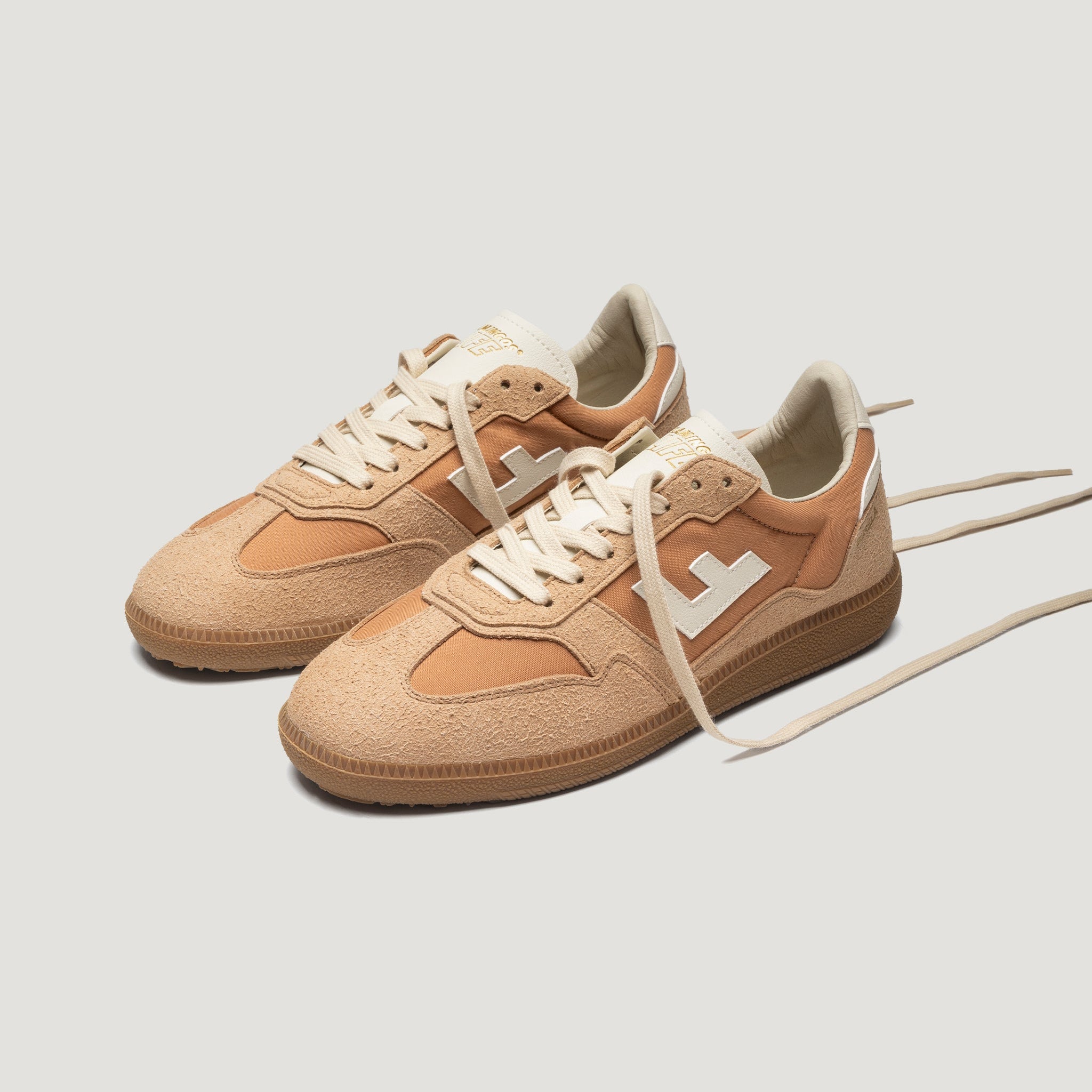 Ein Paar hellbraune Wildleder-Sneakers mit weißen Schnürsenkeln, beigen Details und einem weißen, geometrischen Logo an den Seiten auf hellem Grund.