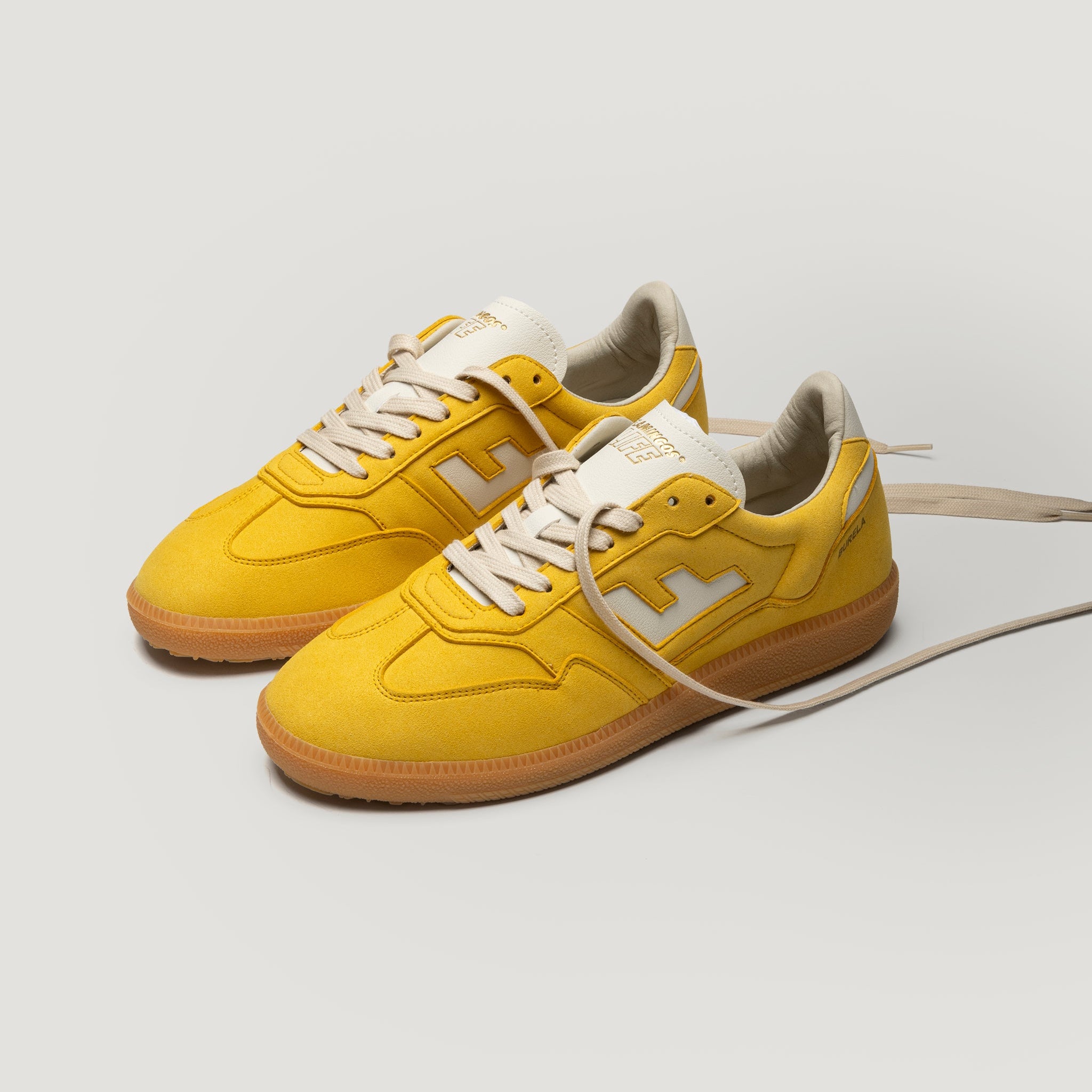 Ein Paar gelbe Turnschuhe mit weißen Schnürsenkeln, beigem Innenfutter und Gummisohlen auf weißem Hintergrund. Ein Schnürsenkel ist vorne herausgezogen.