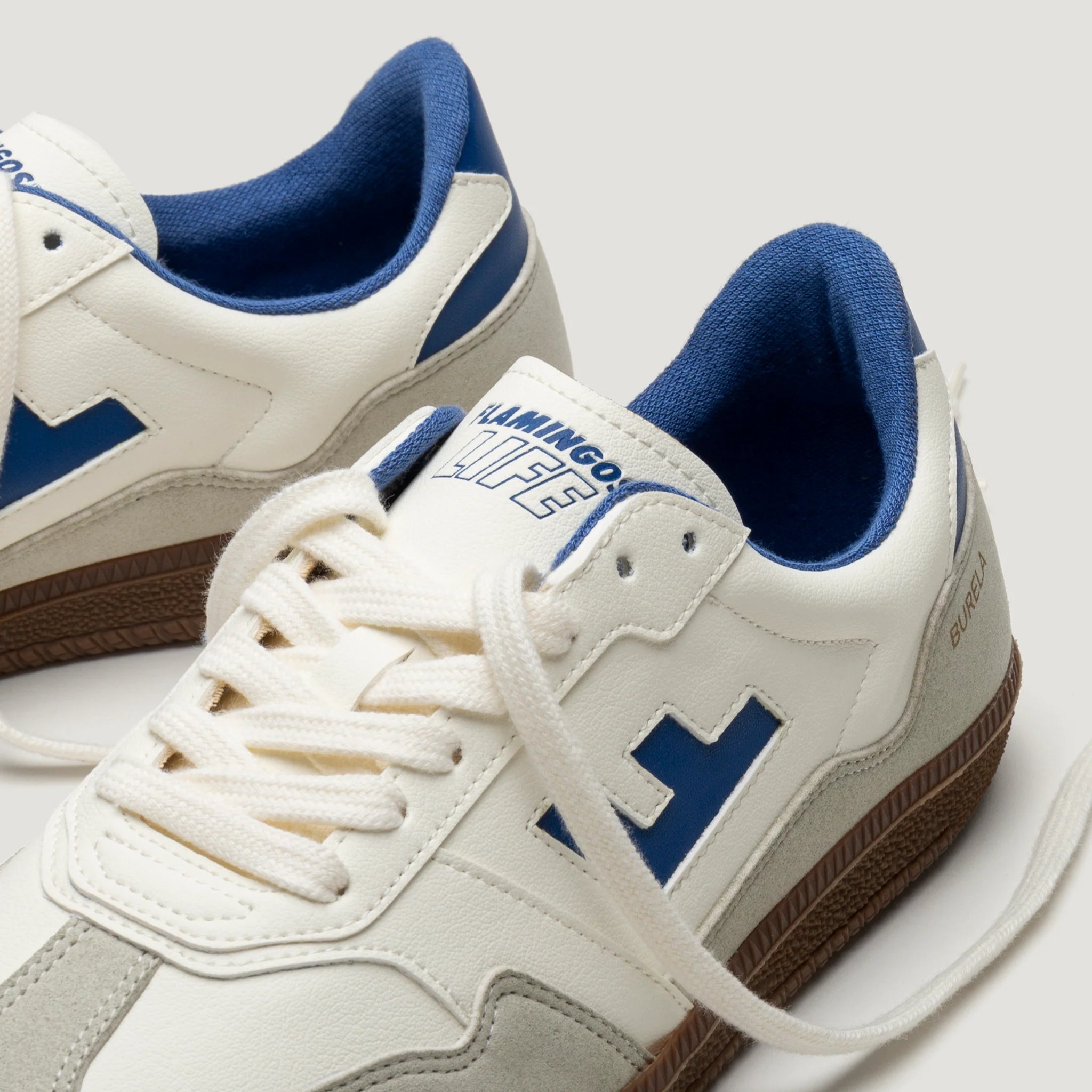 Weiße Turnschuhe mit blauen Details und brauner Sohle auf hellem Hintergrund.