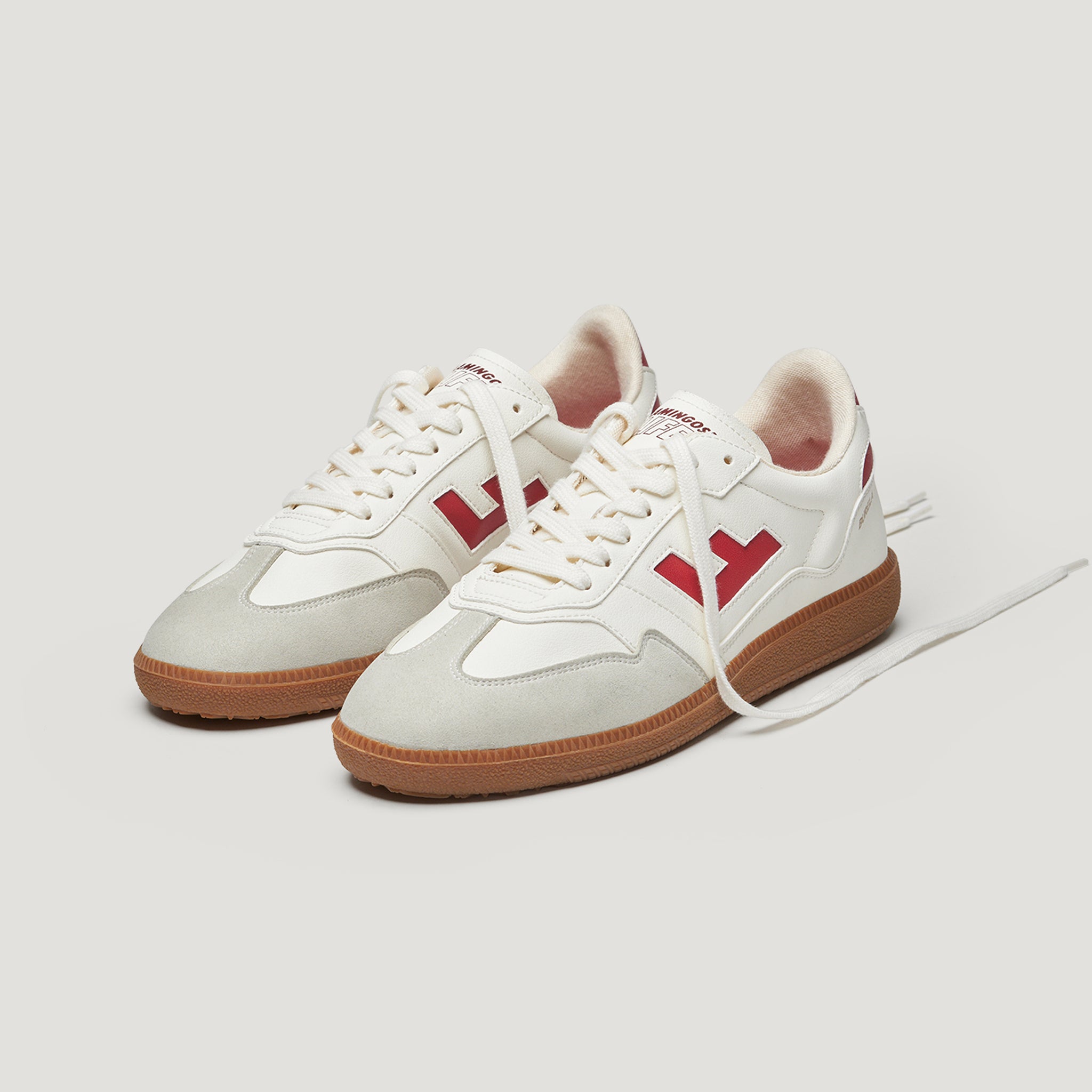 Weiße Sneaker mit roten Logo-Akzenten und brauner Sohle vor grauem Hintergrund.