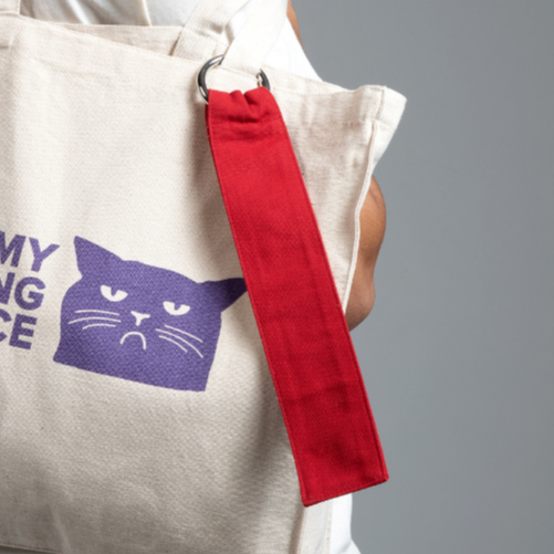Beige Stofftasche mit lila Katzenzeichnung und rotem Griff auf grauem Hintergrund.