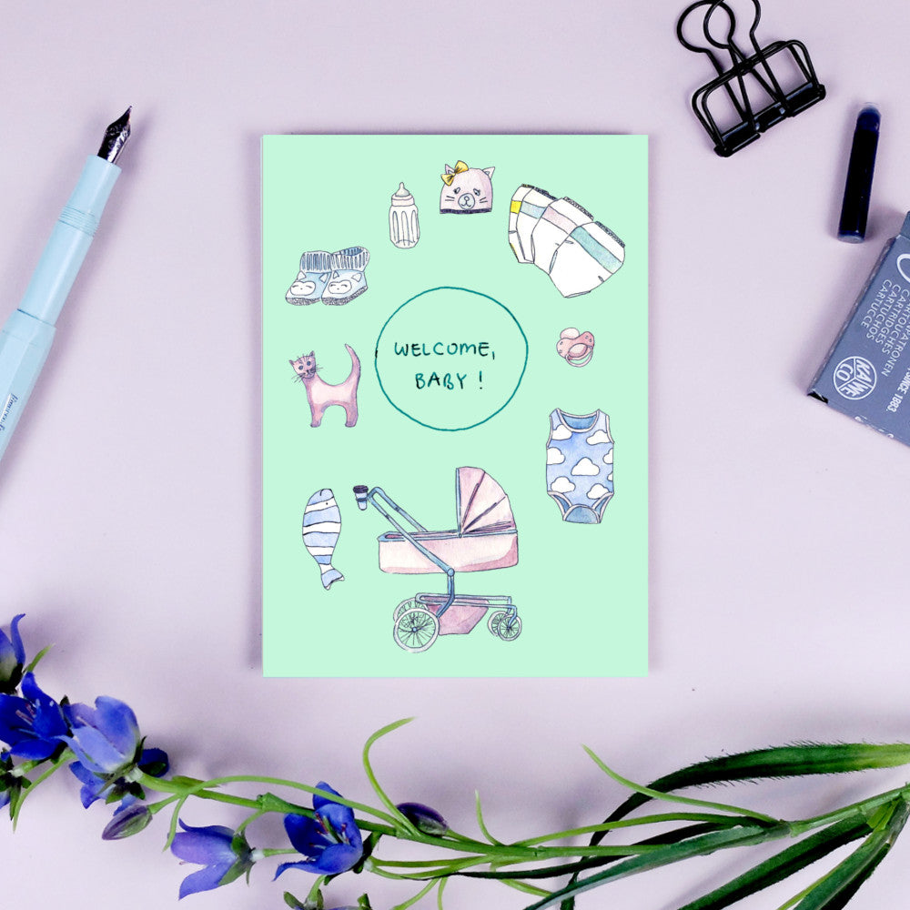 Grüne Karte mit Babysachen-Illustrationen und "Welcome, Baby!"-Text, umgeben von Schreibutensilien und Blumen.