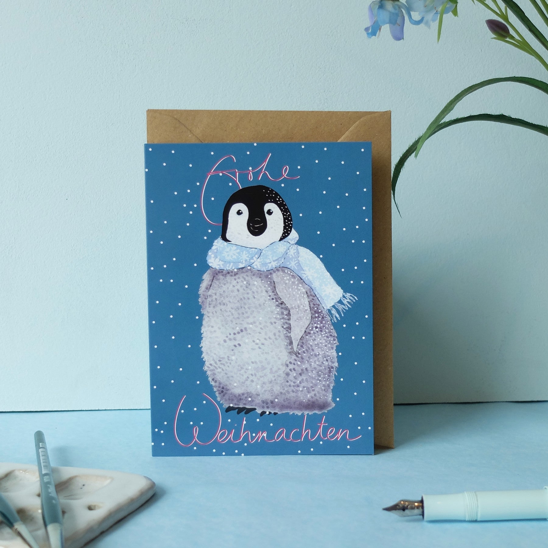 Eine Grußkarte mit einer Illustration eines Pinguinkükens, das einen blauen Schal trägt und auf einem blauen Hintergrund mit weißen Punkten steht. Auf der Karte steht Frohe Weihnachten in rosa Schrift. Ein Umschlag, ein Stift und eine Pflanze liegen daneben.