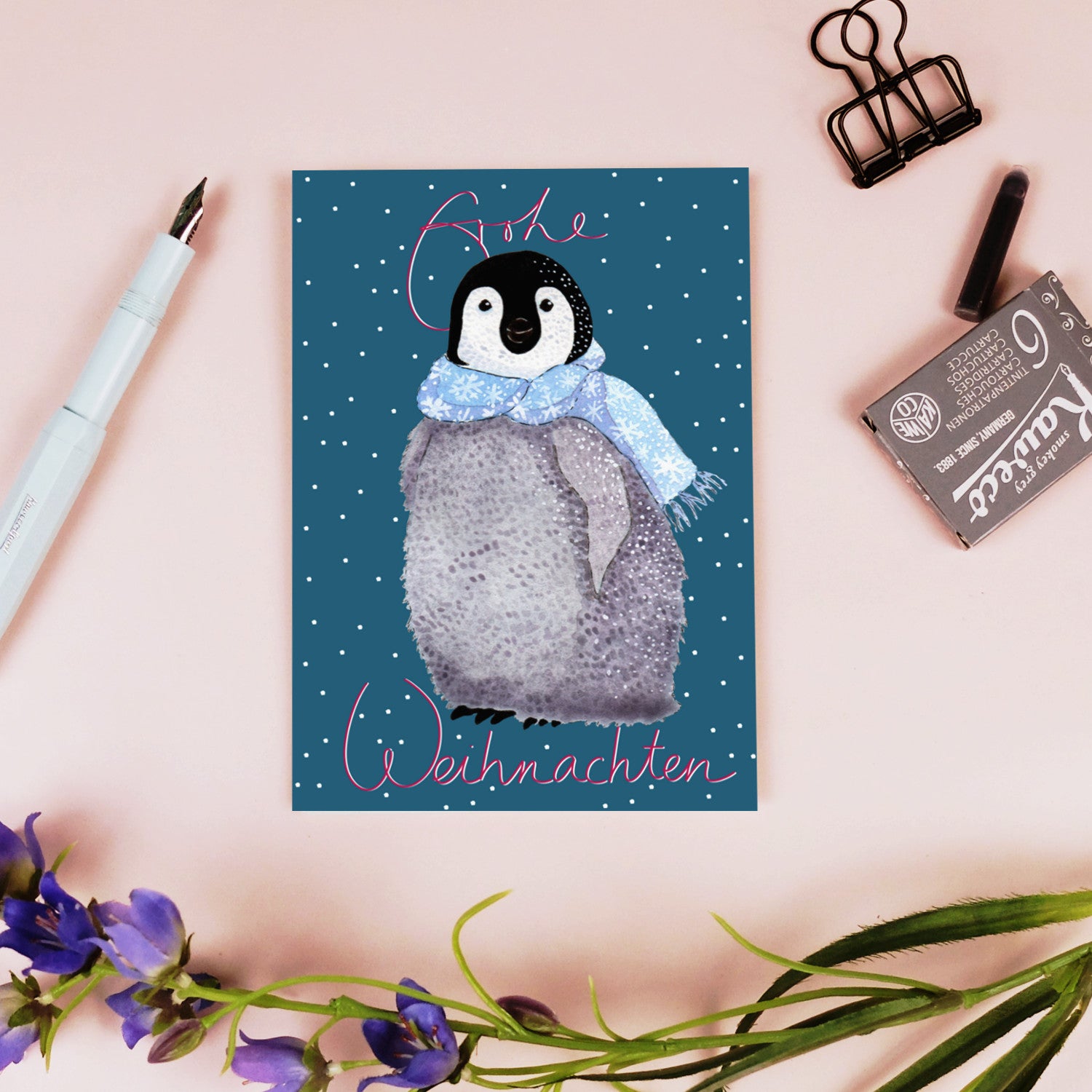 Eine Postkarte mit einem lächelnden Baby-Pinguin, der einen blauen Schal trägt und im fallenden Schnee steht, mit den Worten "Frohe Weihnachten" in rosa geschrieben. Die Karte ist auf einer rosa Oberfläche mit Blumen, einem Stift und Schreibwaren in der Nähe.
