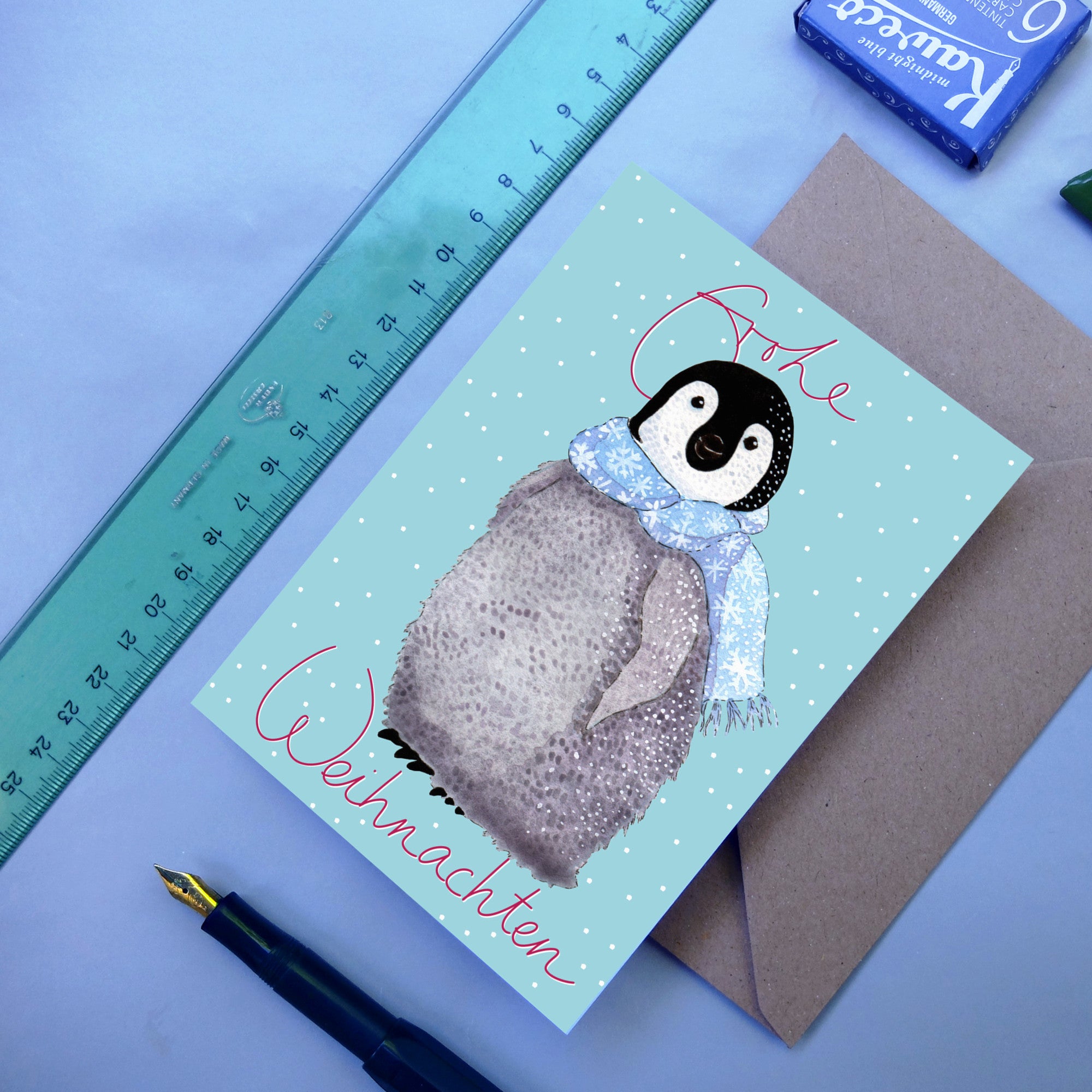 Eine Grußkarte mit einem Pinguinküken, das einen blauen Schal mit Schneeflocken trägt, auf einem hellblauen, verschneiten Hintergrund. Auf der Karte steht Frohe Weihnachten in roter Schrift. In der Nähe befinden sich ein blaues Lineal, ein Stift und ein Umschlag.
