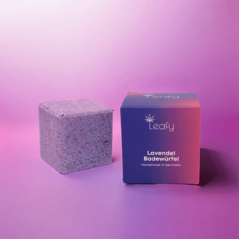 Ein lavendelfarbener Badebombenwürfel sitzt neben einer passenden Farbverlaufsdose mit der Aufschrift Leafy Lavendel Badewürfel Handmade in Germany auf einem rosa und lila Hintergrund.