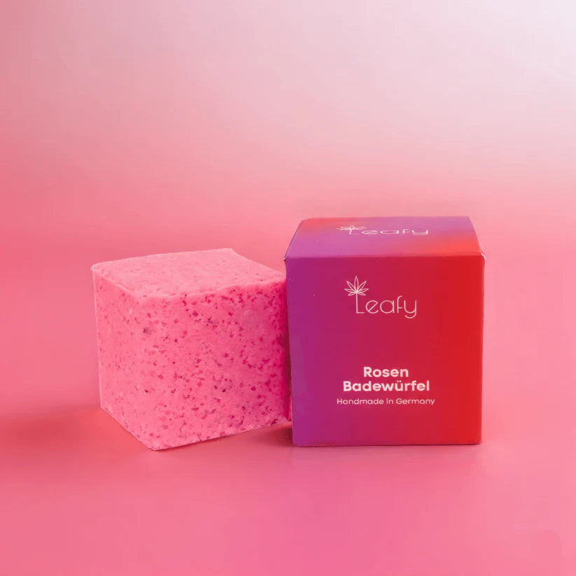 Ein rosafarbener Badewürfel steht neben der dazugehörigen rosafarbenen und lilafarbenen Schachtel mit der Aufschrift "Leafy Rosen Badewürfel Handmade in Germany" vor einem rosa Hintergrund mit Farbverlauf.