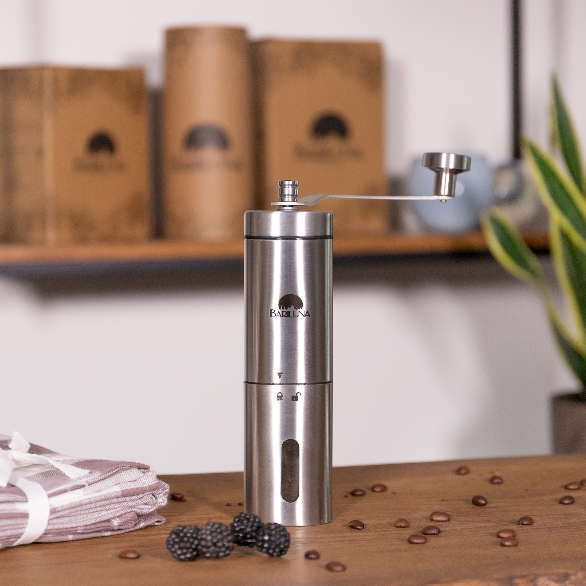 Silberne Kaffeemühle auf einem Holztisch, umgeben von Brombeeren, Kaffeebohnen und gefalteten Stoffservietten.
