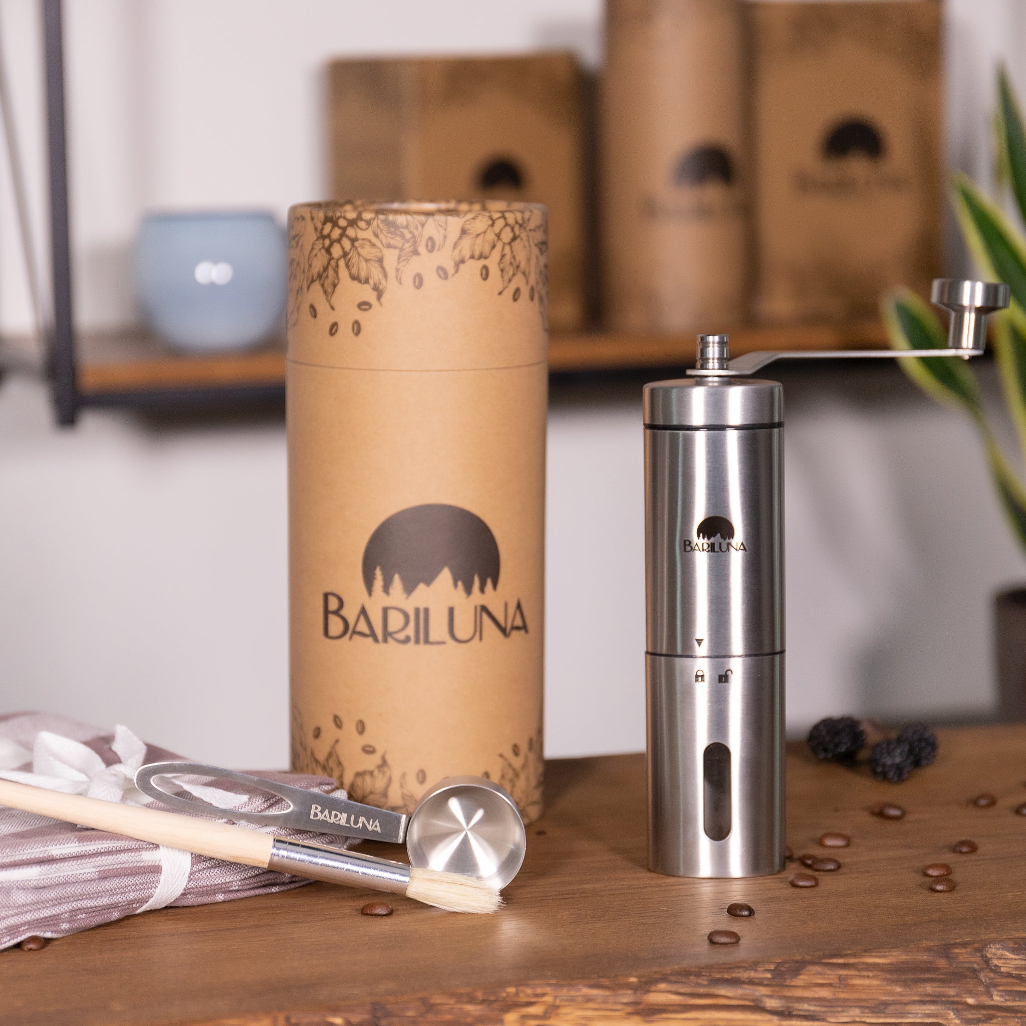 Manuelle Kaffeemühle aus Edelstahl von Bariluna auf einem Holztisch, umgeben von Verpackung, Bürste und Kaffeebohnen.