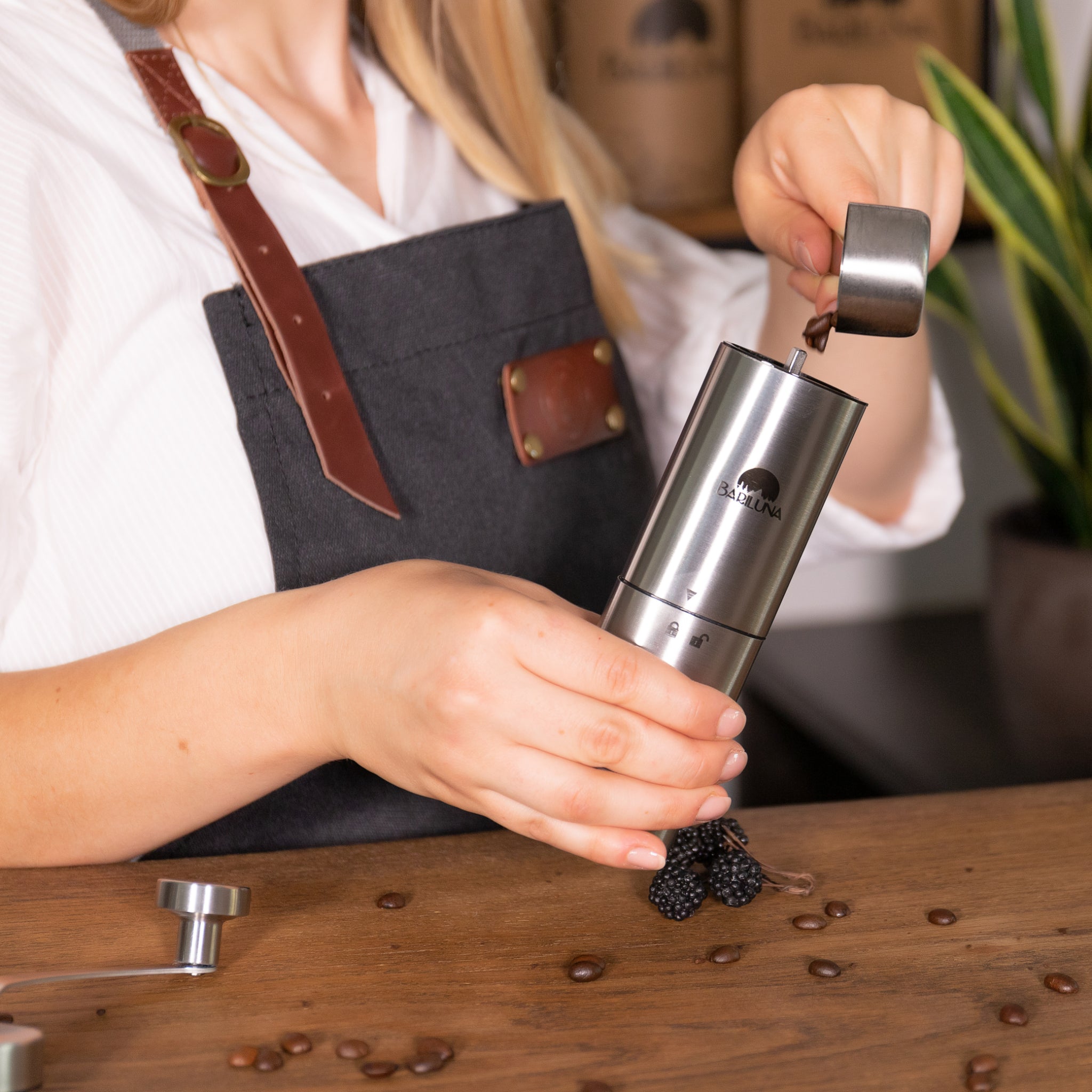 Person mahlt mit einer Handmühle Kaffeebohnen, auf einem Holztisch liegen Brombeeren und Kaffeebohnen verstreut.