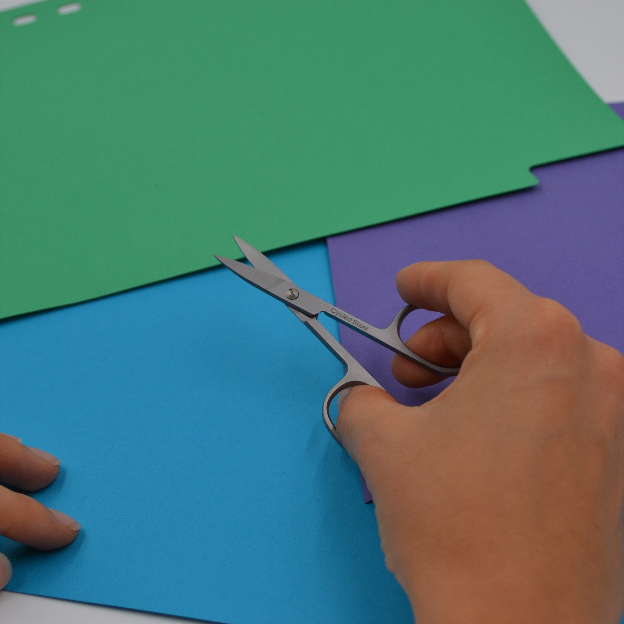 Eine Person hält eine kleine Schere in der Hand und bereitet sich darauf vor, ein Blatt blaues Papier zu schneiden. In der Nähe liegen Blätter mit grünem und violettem Papier auf einer ebenen Fläche.