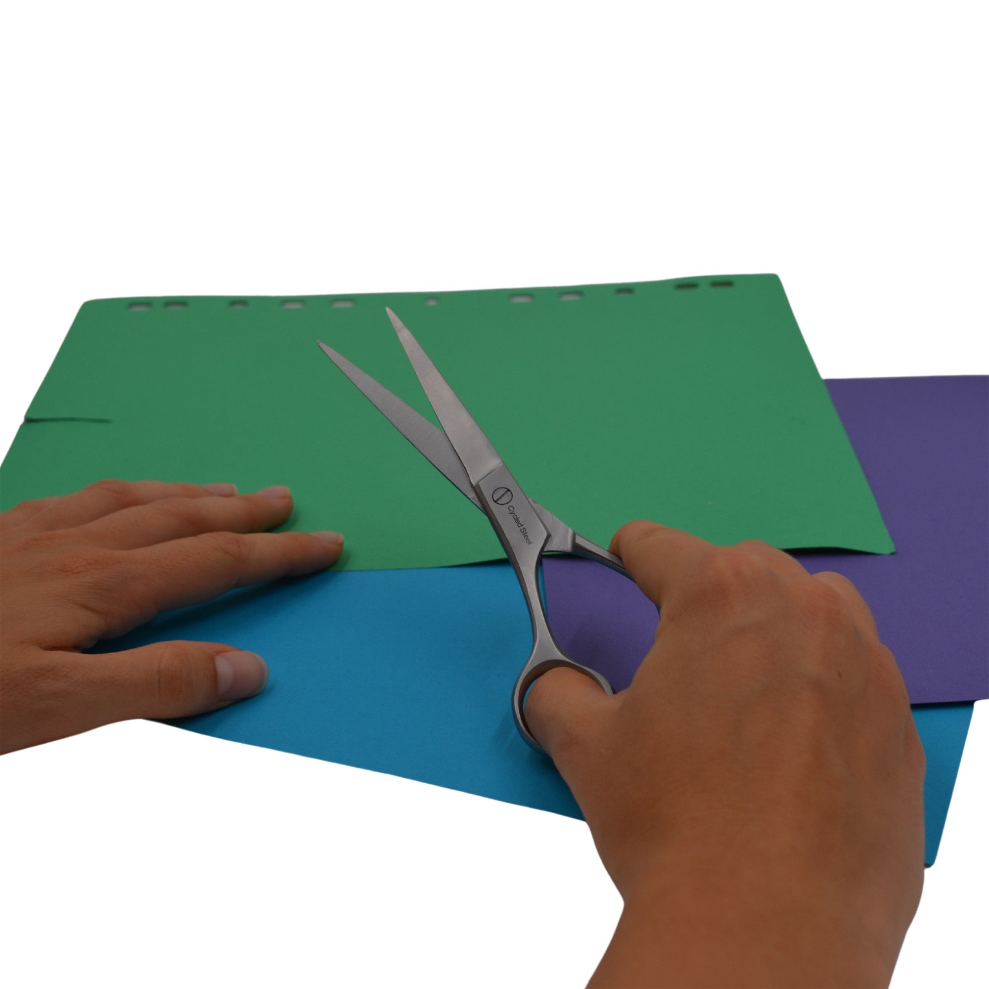 Die Hände einer Person, die eine Schere hält, schneiden durch blaues Papier, das auf grünen und lila Blättern liegt, vor einem weißen Hintergrund.