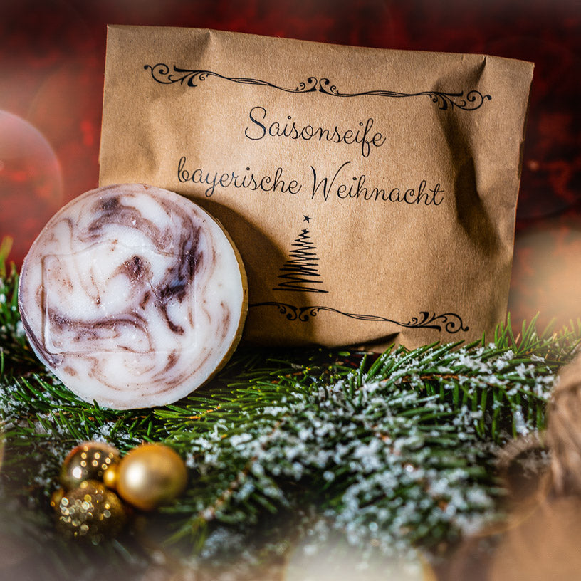 Rundes Seifenstück und braune Verpackung mit der Aufschrift "Saisonseife bayerische Weihnacht" auf Tannenzweigen.