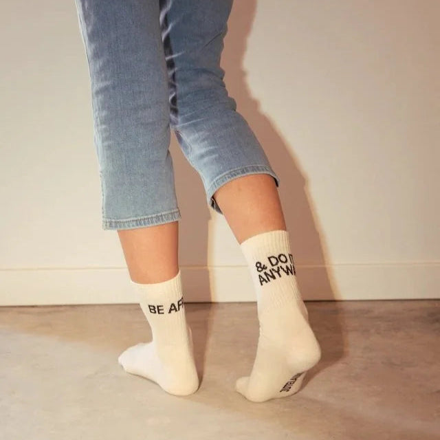 Eine Person in hellblauen Jeans und weißen Socken steht auf Zehenspitzen. Die Socken tragen die Aufschrift: BE AFRAID auf der einen und & DO IT ANYWAY auf der anderen. Der Hintergrund ist eine schlichte Wand und der Boden.