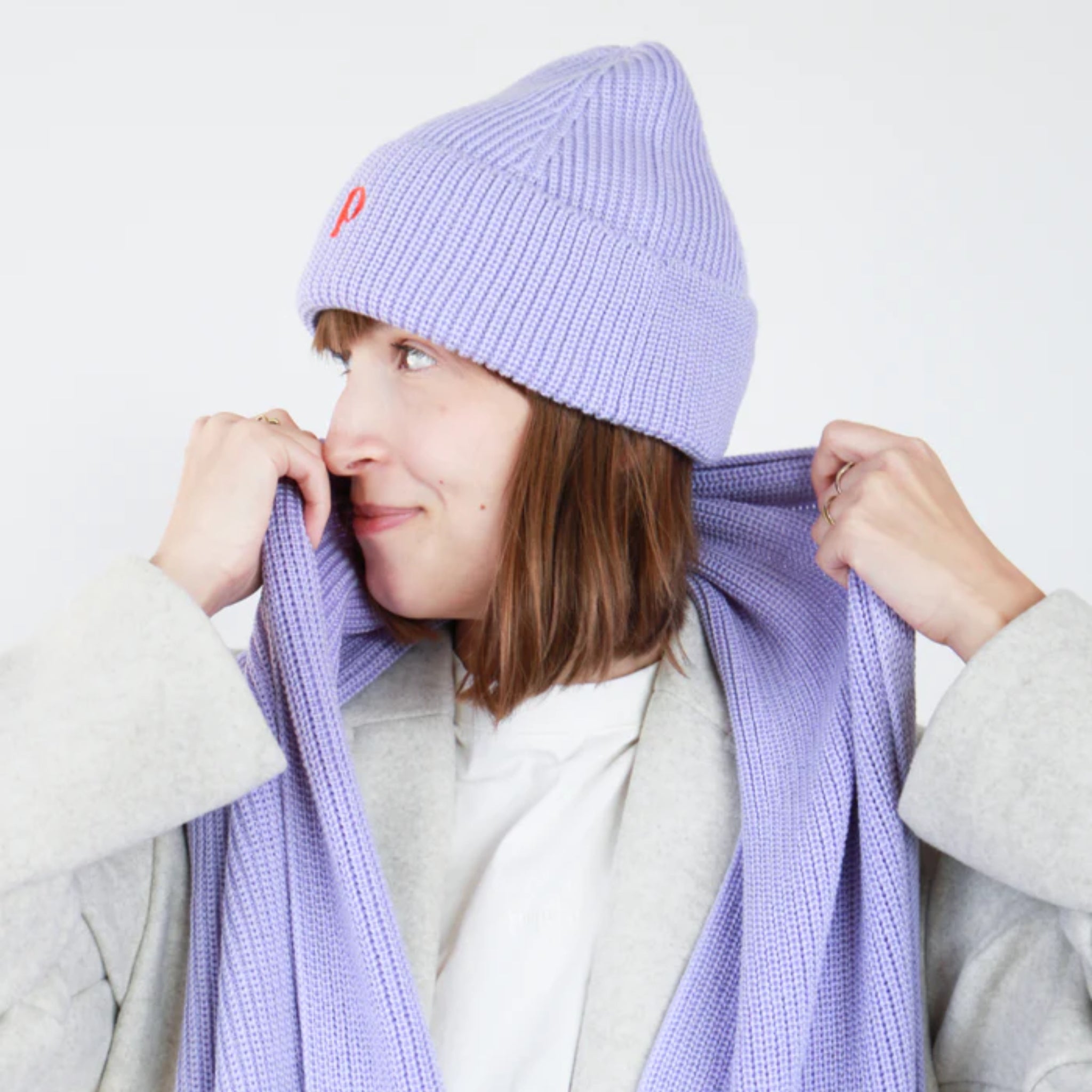 Frau in grauem Mantel und lila Mütze passt einen passenden Schal an und lächelt seitlich in die Kamera.
