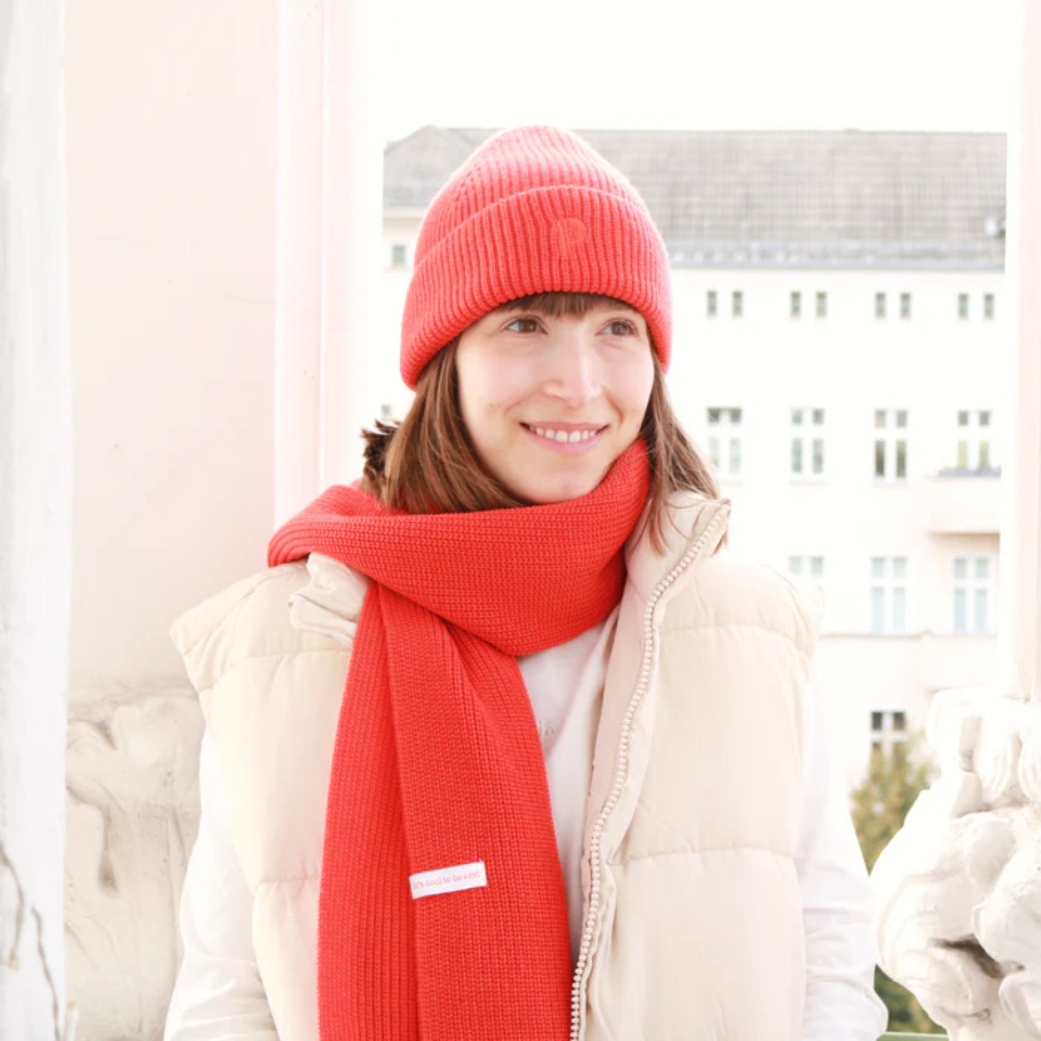 Frau in beige Weste mit roter Mütze und Schal steht draußen vor einem Gebäude und lächelt.