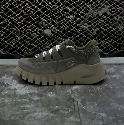 Grüner Sneaker mit dicker Sohle und weißen Schnürsenkeln auf grauem Boden vor einem Maschendrahtzaun.