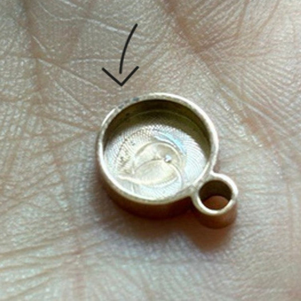 Ein runder, goldfarbener Anhänger mit einer kleinen Schlaufe sitzt auf einer Handfläche einer Person. Ein Pfeil zeigt auf die leere, flache Mitte des Anhängers, was darauf hindeutet, dass etwas fehlt oder im Inneren platziert werden soll.