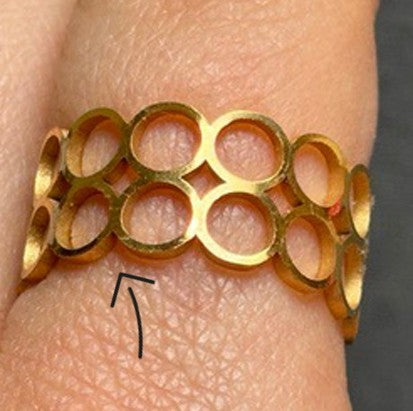 Nahaufnahme eines Goldrings mit einem offenen Kreismuster an einem Finger. Ein schwarzer Pfeil zeigt auf eine kreisförmige Vertiefung, die das Ringdesign auf der Haut hinterlassen hat.