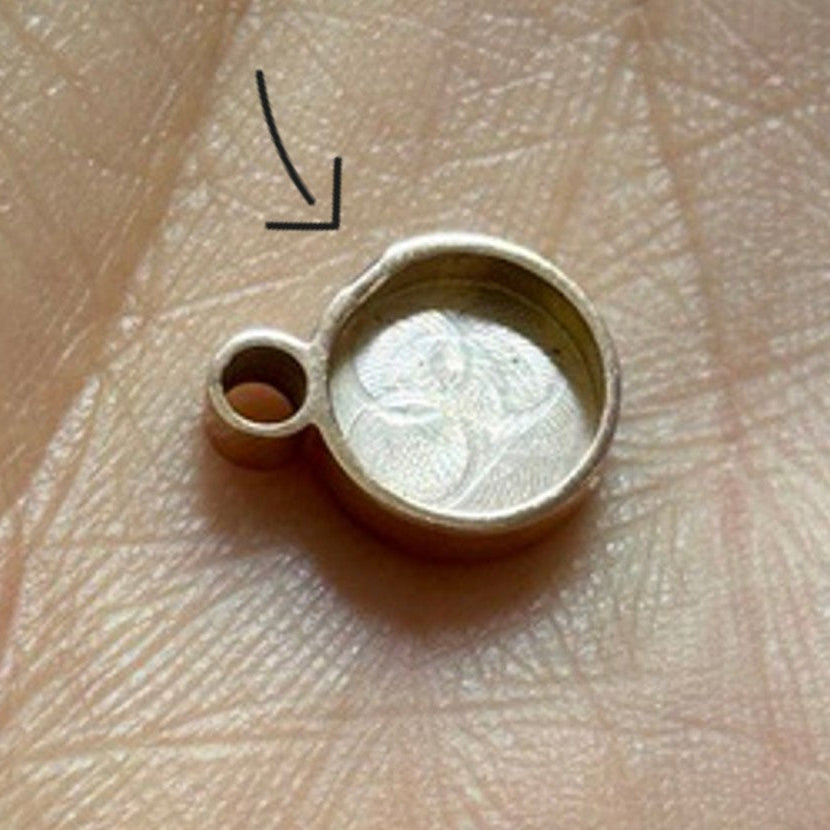 Ein kleiner, runder, goldfarbener Metallanhänger mit erhöhtem Rand und einer Schlaufe zum Aufhängen ruht auf einer Handfläche. Darüber ist ein Pfeil eingezeichnet, der auf eine flache Kerbe am Rand des Anhängers zeigt.
