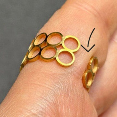 Ein Goldring mit mehreren offenen Kreisen wird an einem Finger getragen. Ein Pfeil zeigt auf einen Teil des Rings und hebt drei der Kreisformen hervor. Der Hintergrund ist ein verschwommenes Grau.