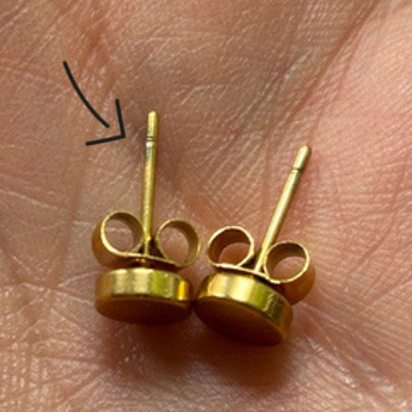Nahaufnahme einer Hand, die zwei goldene Ohrstecker hält, mit einem schwarzen Pfeil, der auf eine schwache Linie in der Nähe des oberen Endes des einen Ohrsteckers zeigt.