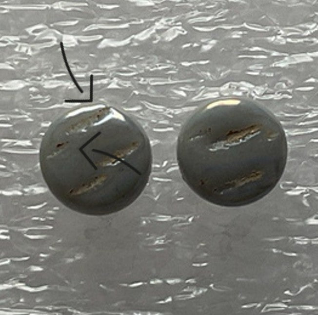 Zwei runde, graue Knöpfe auf einer strukturierten Oberfläche. Der linke Knopf hat drei sichtbare Kratzspuren und ist mit schwarzen Pfeilen markiert, die auf die Kratzer zeigen.