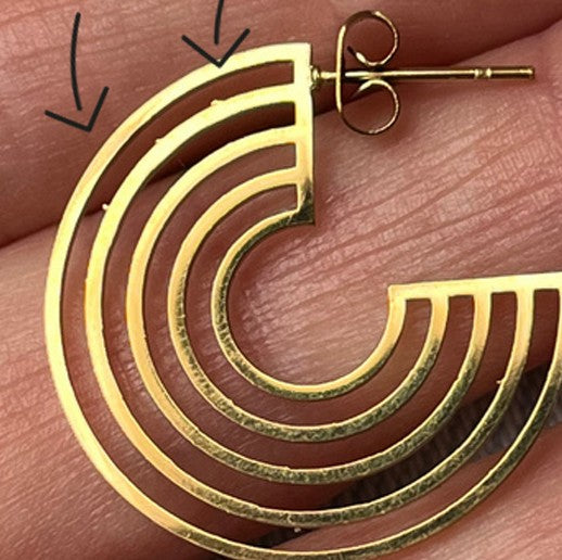 Nahaufnahme einer Hand, die einen goldenen, geometrischen Ohrring in Form eines unvollständigen Kreises mit parallelen Linien hält. Auf dem Bild sind schwarze Pfeile eingezeichnet, die auf den äußersten Teil des Ohrrings zeigen.