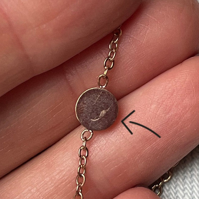 Nahaufnahme einer Hand, die ein goldenes Kettenarmband mit einem runden, braunen Anhänger hält. Ein Pfeil zeigt auf eine schwache, weiße Markierung auf der Oberfläche des Anhängers.