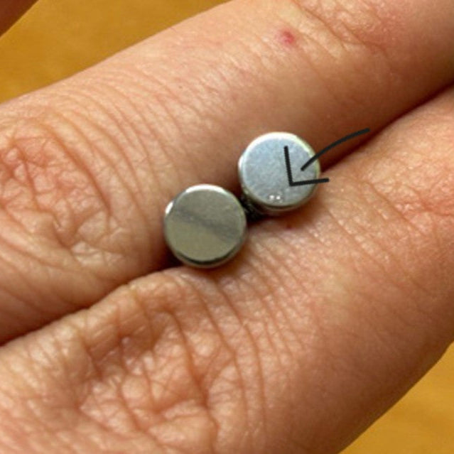 Eine Nahaufnahme von zwei kleinen runden Metallbatterien, die auf den Fingern einer Person liegen. Ein Pfeil zeigt auf eine kaum sichtbare Beule oder Markierung auf der Oberfläche der einen Batterie.