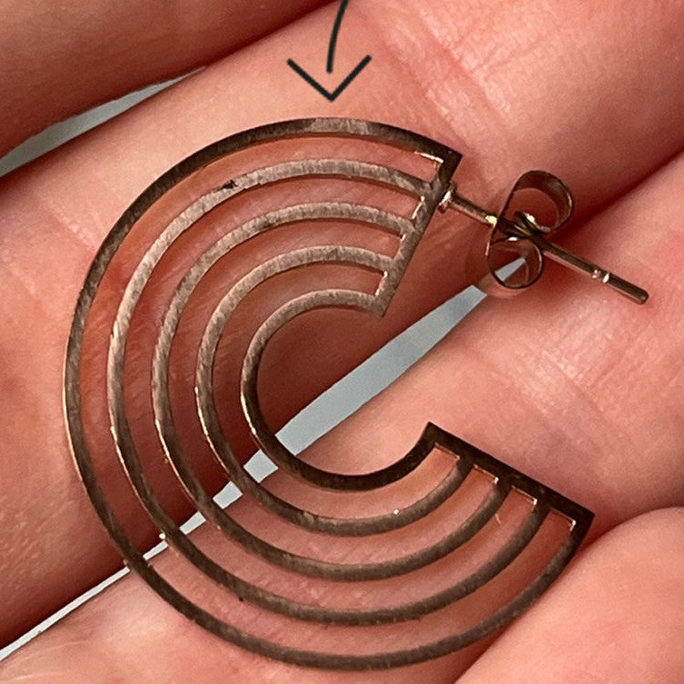 Eine Hand hält einen metallischen Ohrring, der die Form eines geometrischen, offenen Halbkreises mit drei parallelen Bögen hat. Darüber ist ein Pfeil eingezeichnet, der auf den Ohrring zeigt.