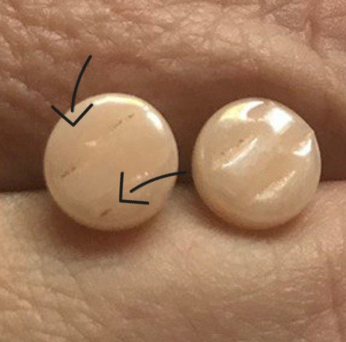 Zwei runde, glatte, hellbeige Steine sind auf einer strukturierten Oberfläche platziert. Beide haben schwache Linien, und schwarze Pfeile zeigen auf diese Linien, die möglicherweise Unterschiede oder Merkmale der Steine hervorheben.