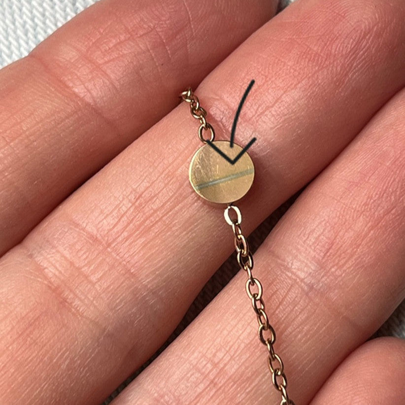 Nahaufnahme einer Hand, die ein goldenes Kettenarmband mit einem kleinen ovalen Anhänger hält. Auf dem Bild ist ein schwarzer Pfeil eingezeichnet, der auf das Schildchen zeigt, das eine schwache Gravur aufweist.