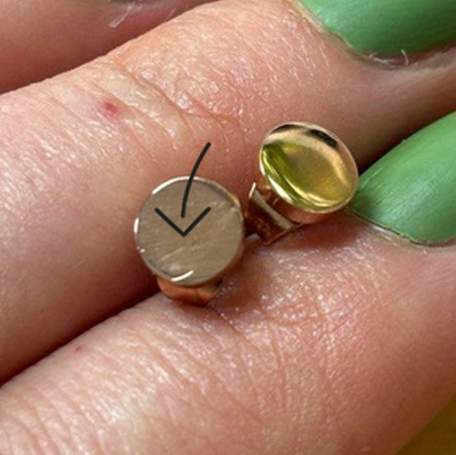 Eine Nahaufnahme von zwei Fingern mit grünem Nagellack, die einen goldenen Ohrring halten, der mit einem schwarzen Pfeil markiert ist, um einen kleinen Kratzer oder eine Vertiefung auf der Oberfläche hervorzuheben.