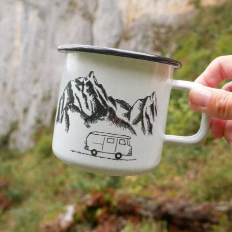 Eine Hand hält einen weißen Metallbecher mit schwarzem Rand, auf dem eine Illustration von Bergen und einem Lieferwagen zu sehen ist. Der Hintergrund ist unscharf mit Grün und einer felsigen Klippe.