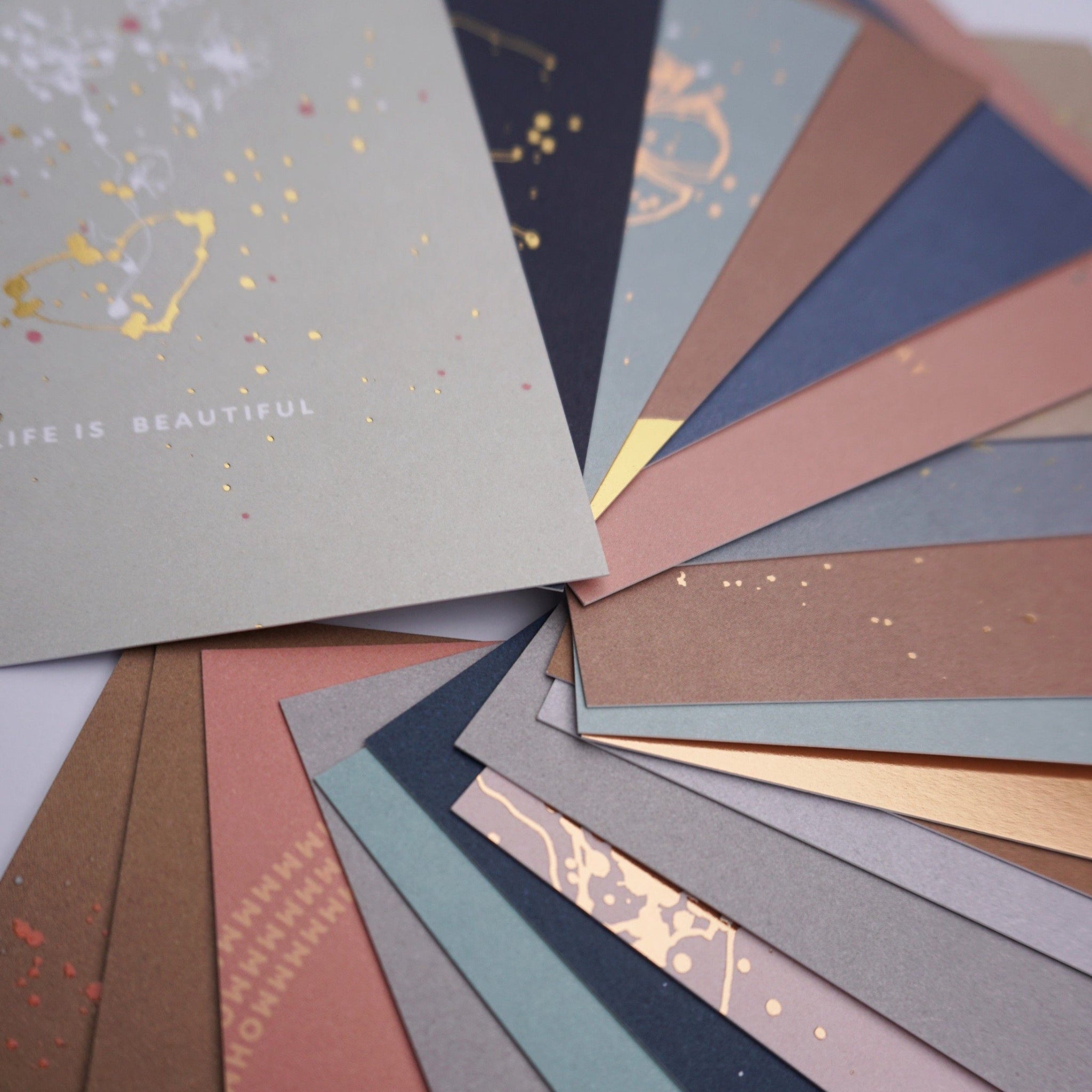 Fächer aus buntem Papier mit goldenen Sprenkeln, einem Blatt mit der Aufschrift "LIFE IS BEAUTIFUL" im Vordergrund.