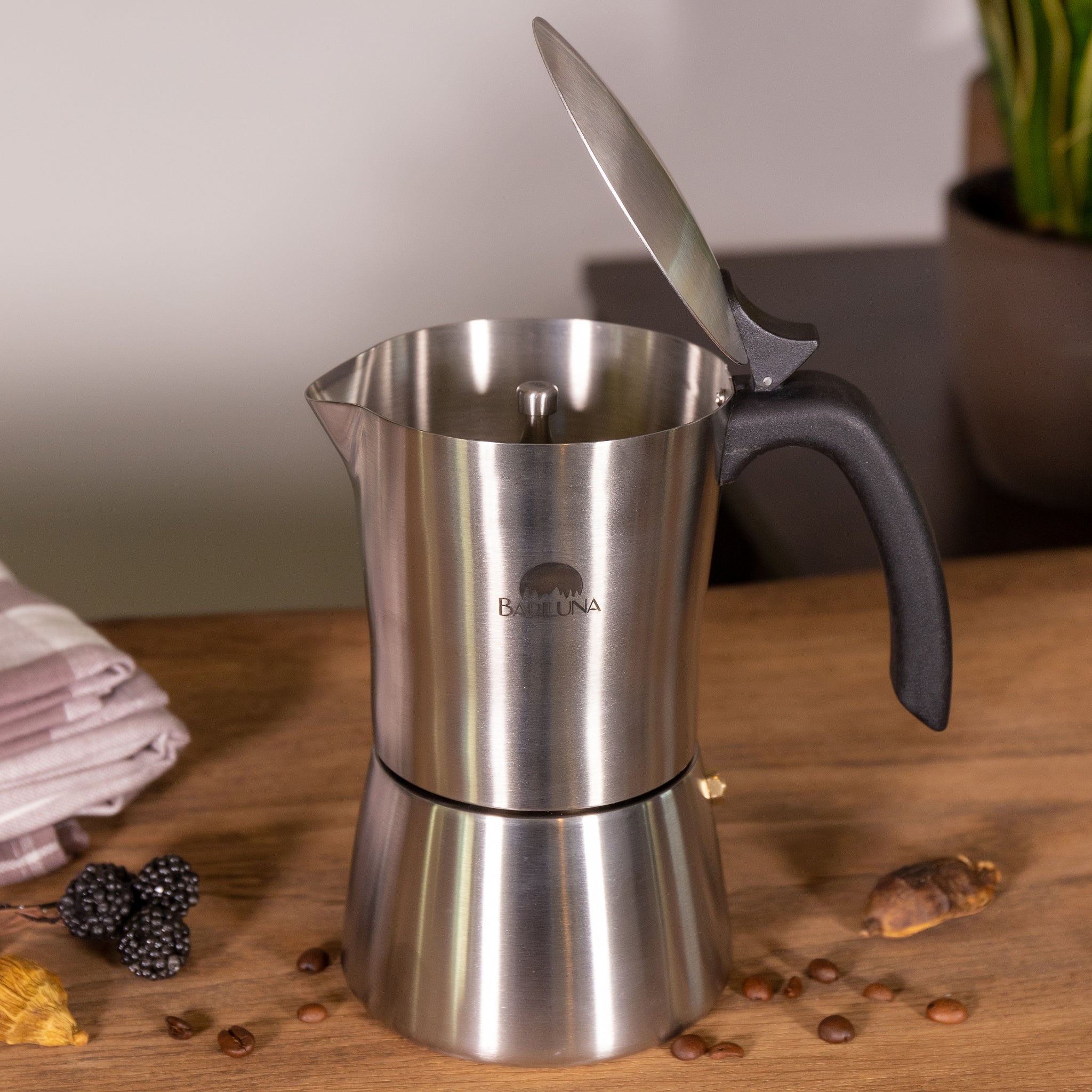 Edelstahl-Espressokocher mit offenem Deckel auf einem Holztisch, umgeben von Kaffeebohnen und Brombeeren.