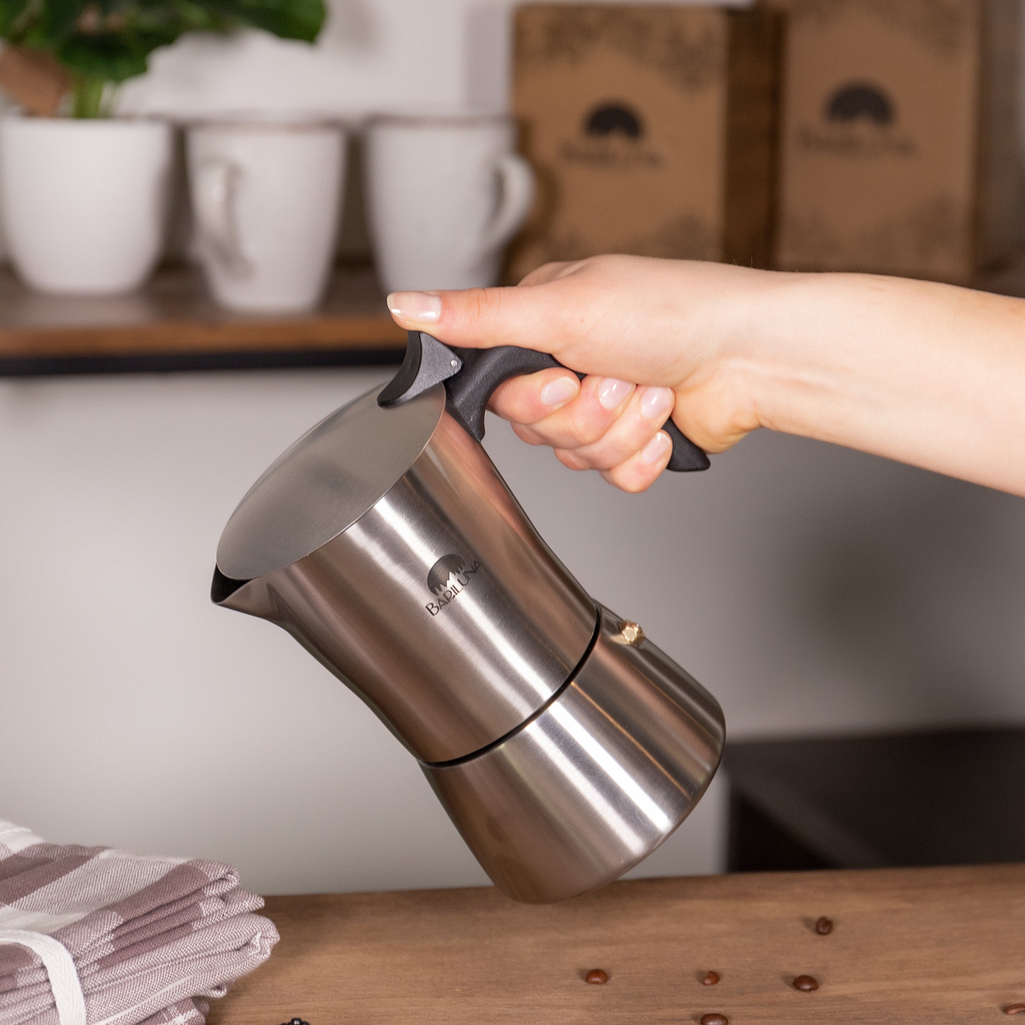 Eine Hand hält eine silberne Espressokanne über einem Tisch mit Kaffeebohnen. Im Hintergrund stehen Tassen.