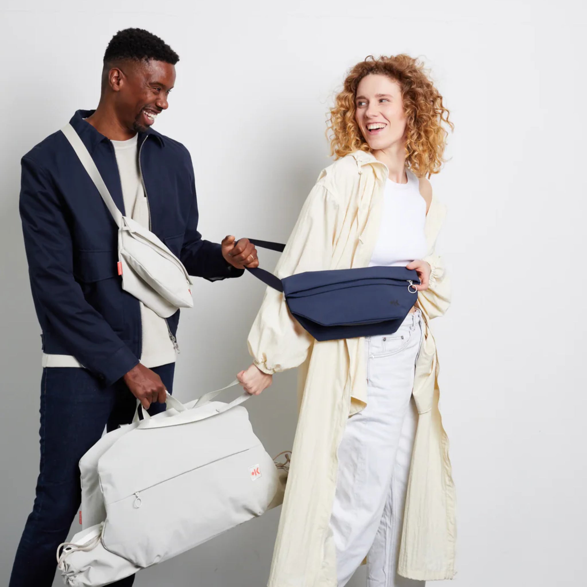 Ein Mann und eine Frau lächeln sich an und halten sich an den Händen. Der Mann trägt eine marineblaue Jacke, ein schwarzes Hemd und hat eine graue Umhängetasche und einen Seesack dabei. Die Frau trägt ein helles Kleid und eine blaue Gürteltasche vor der Brust.