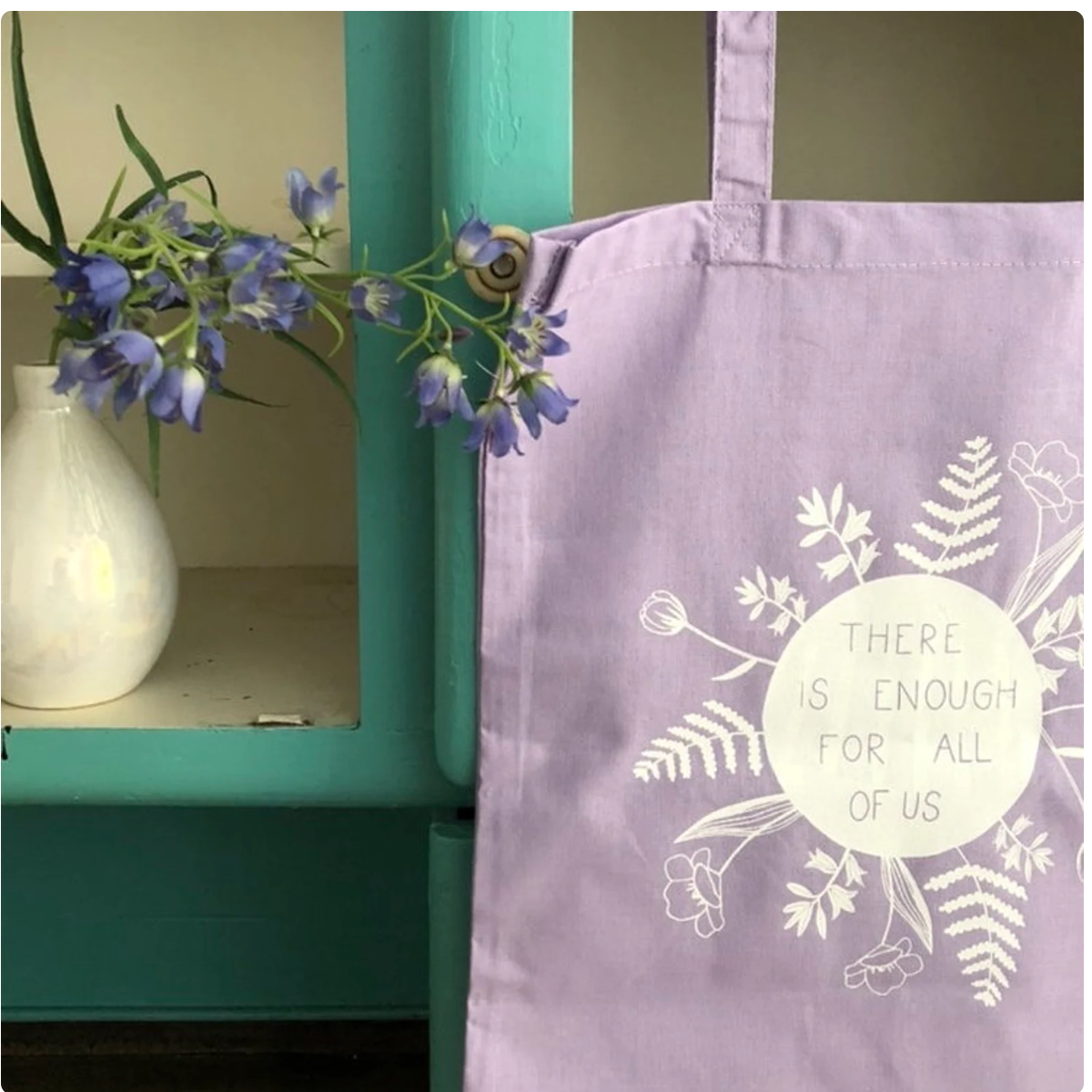 Lilafarbene Tasche mit der Aufschrift „THERE IS ENOUGH FOR ALL OF US“, daneben eine Vase mit lila Blumen.
