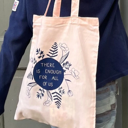 Person trägt eine beigefarbene Stofftasche mit der Aufschrift "There is enough for all of us" und floralen Motiven.
