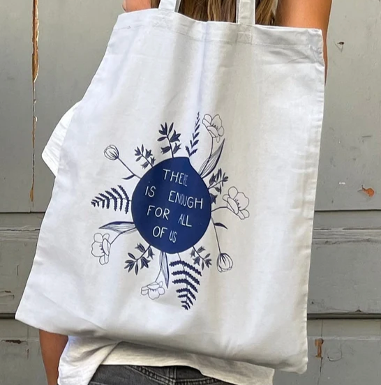 Frau trägt einen Stoffbeutel mit der Aufschrift „There is enough for all of us“ umgeben von Blumenmuster.