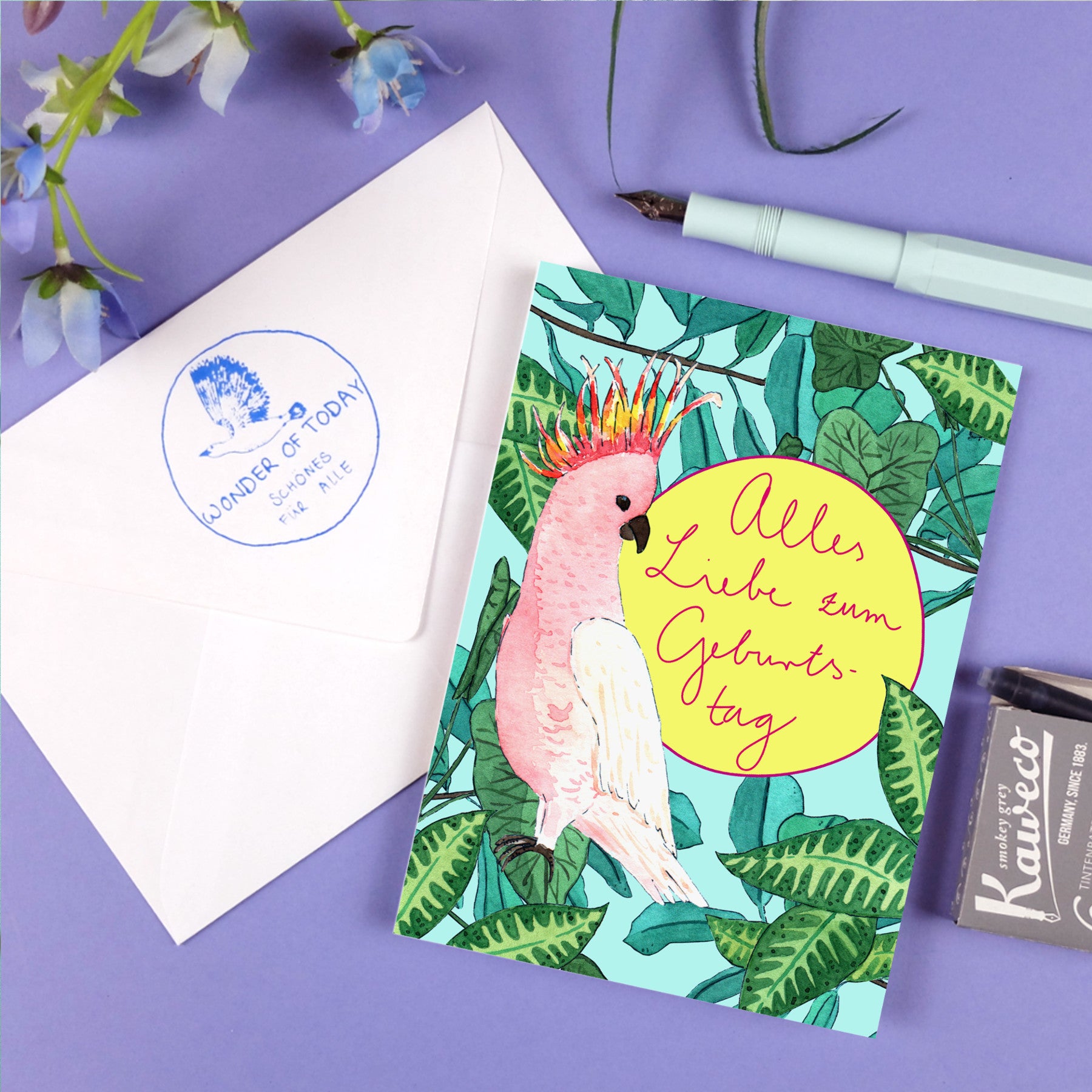 Eine farbenfrohe Geburtstagskarte mit einem rosafarbenen Kakadu und grünen Blättern mit dem deutschen Text Alles Liebe zum Geburtstag liegt neben einem weißen Umschlag, einem hellblauen Stift, einer Blume und einem schwarzen Radiergummi auf einer lila Fläche.