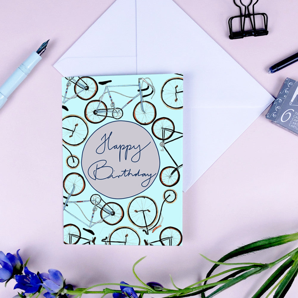 Geburtstagskarte mit Fahrradmuster und der Aufschrift "Happy Birthday" auf rosa Hintergrund, umgeben von Schreibwaren.