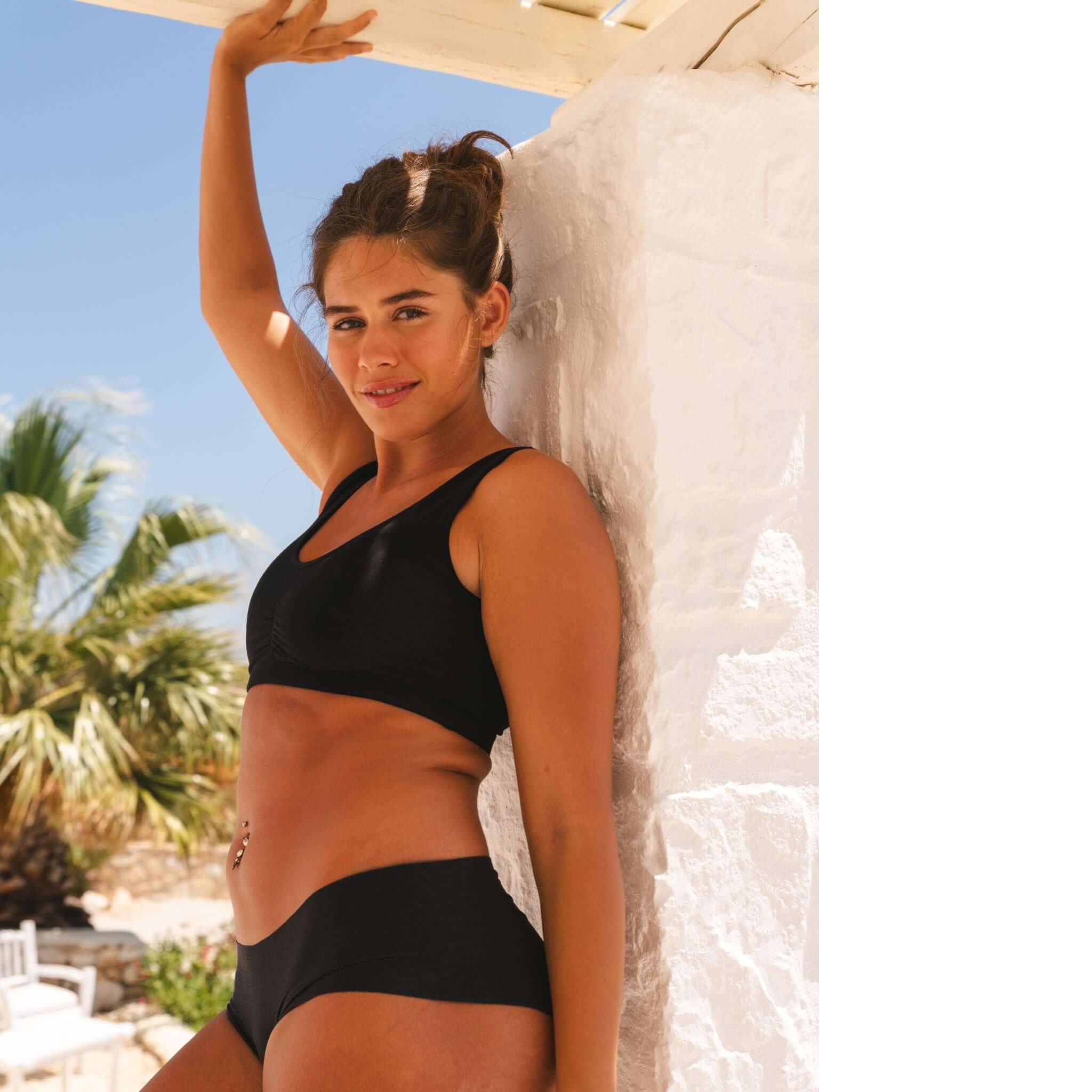 Eine Frau in schwarzem Badeanzug lehnt an einer weißen Wand, mit einem Palmenhintergrund und blauem Himmel.