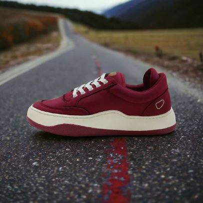 Weinroter Sneaker mit weißen Schnürsenkeln liegt auf einer Landstraße, umgeben von Landschaft mit Bergen im Hintergrund.