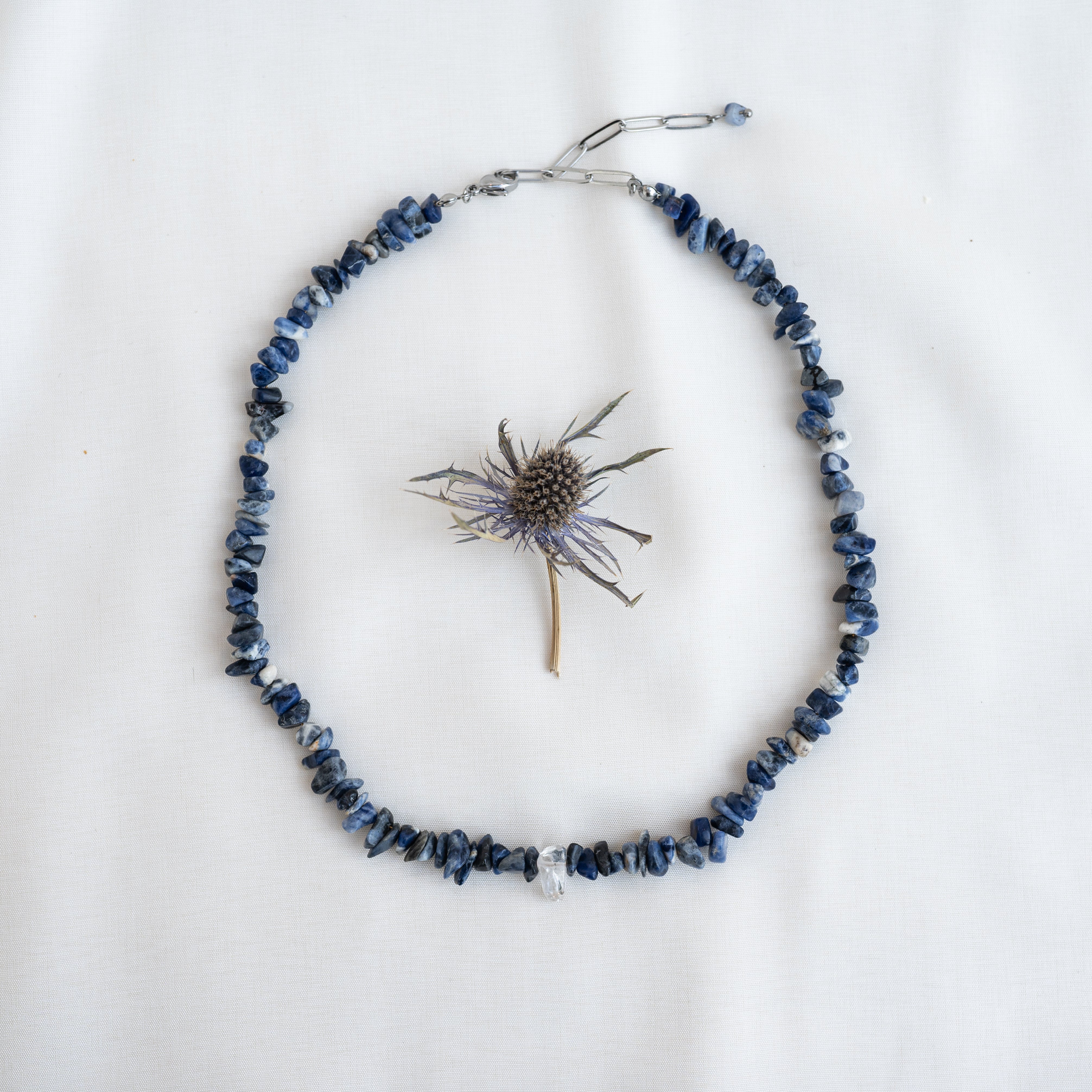 Eine Halskette aus unregelmäßigen blauen Steinperlen ist kreisförmig auf weißem Hintergrund angeordnet, in der Mitte der Kette befindet sich eine getrocknete Distelblüte.