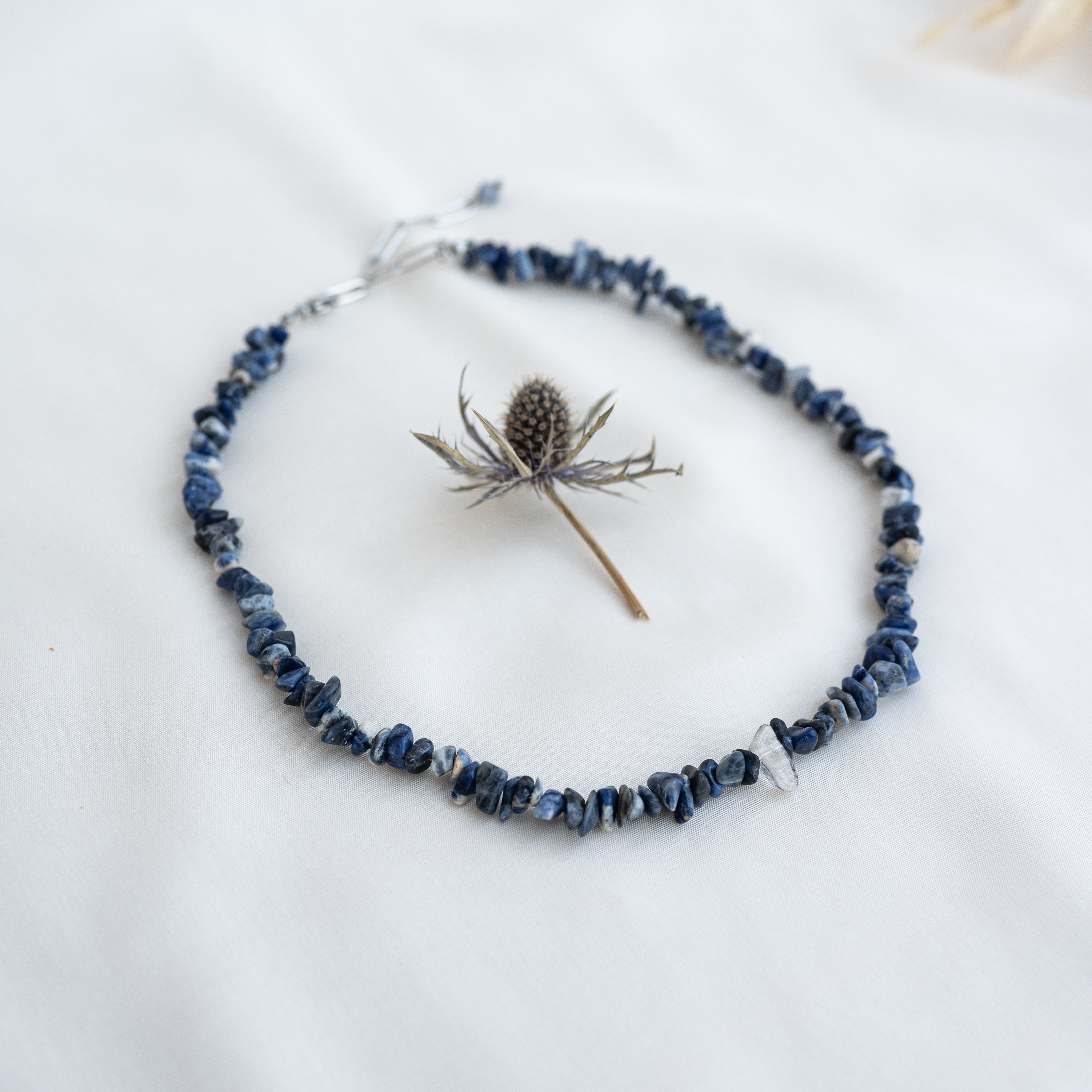 Eine blaue Edelsteinchip-Halskette, kreisförmig angeordnet auf einem weißen Stoffhintergrund, mit einer getrockneten Distelblüte in der Mitte der Kette.