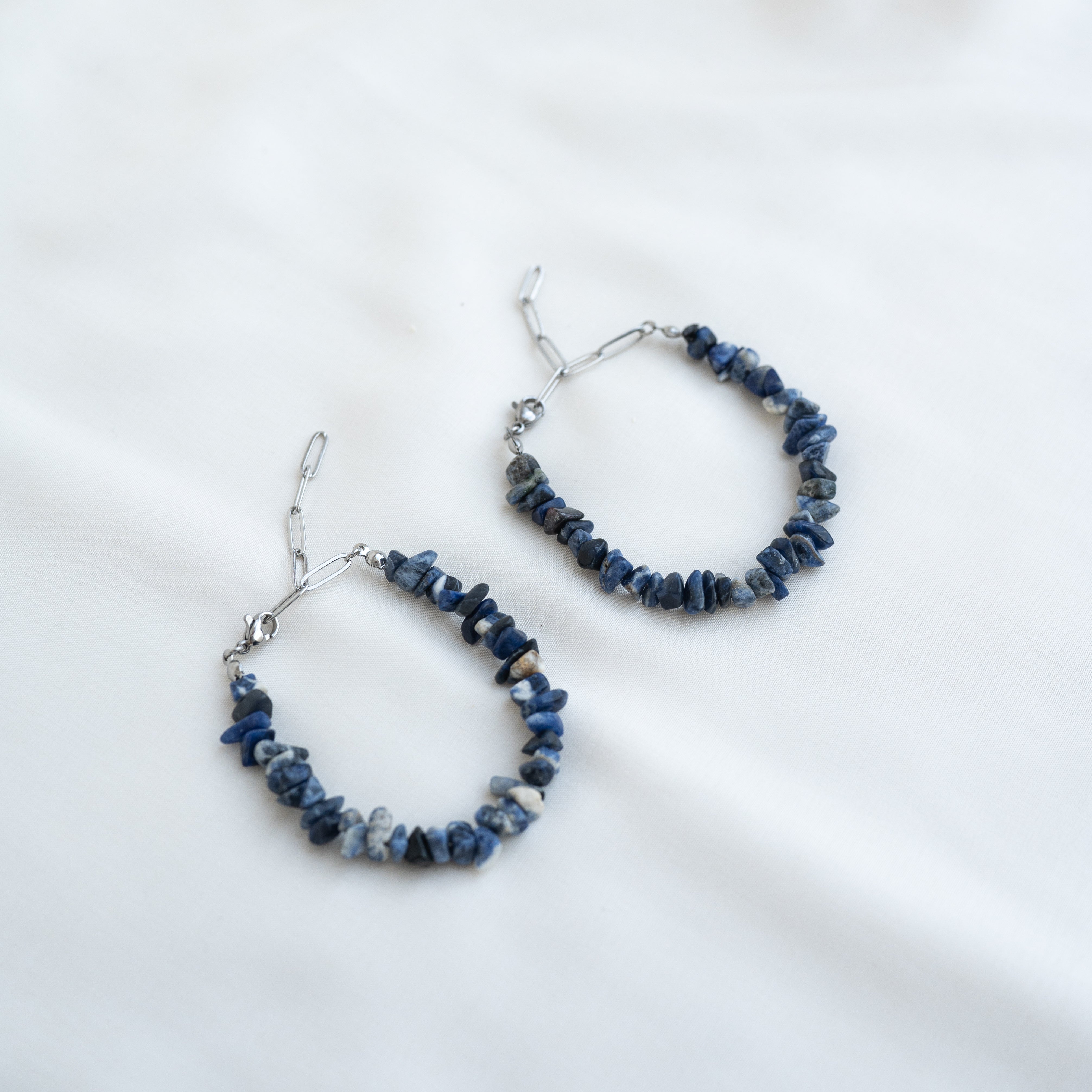 Ein Paar Creolen mit unregelmäßig geformten blauen Steinen und silbernen Drahtverschlüssen auf weißem Stoff.